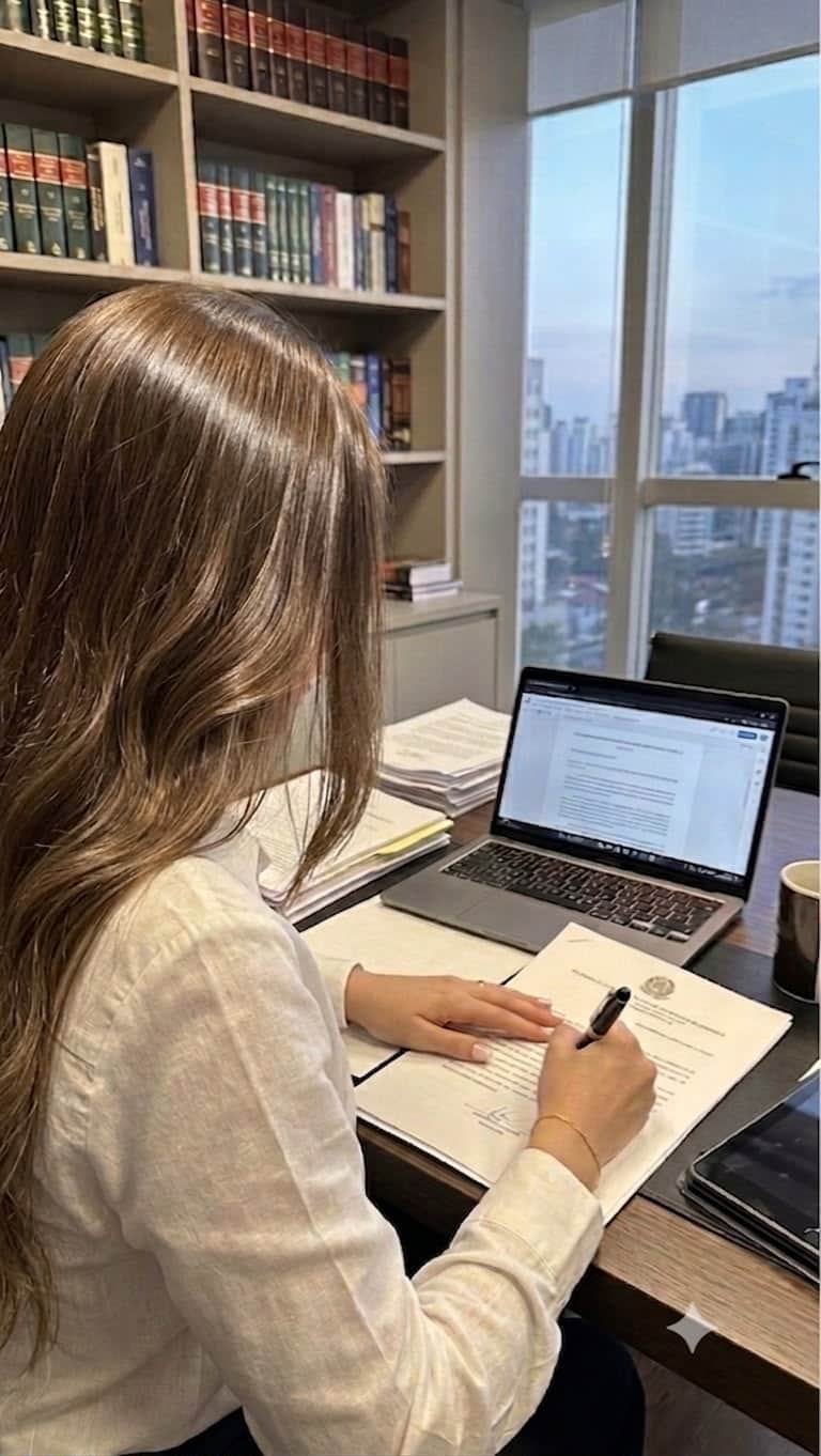 Vitória Mercêz assinando uma peça processual em sua mesa, com autos impressos e o notebook aberto ao lado